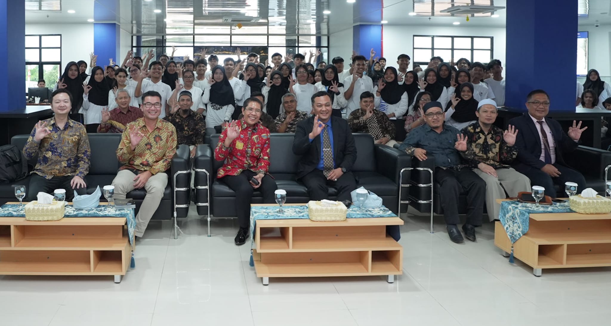 Large group of people in a hall, some seated, waving with okay hand gestures.